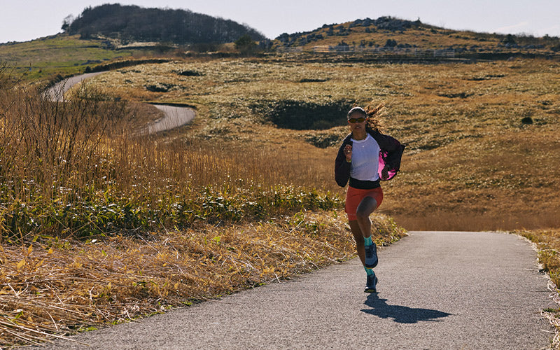 A trail runner wearing a white tank top, pink and black jacket, and orange shorts is running on a paved path through a dry, grassy landscape with rolling hills under a clear sky.