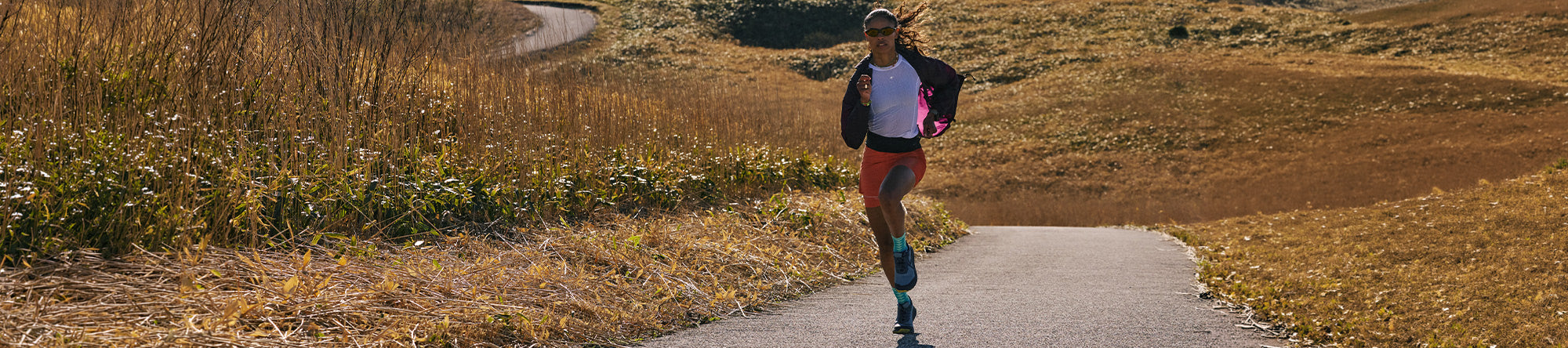A woman running on a paved path through a grassy outdoor landscape, wearing a black jacket, white top, and colorful leggings.